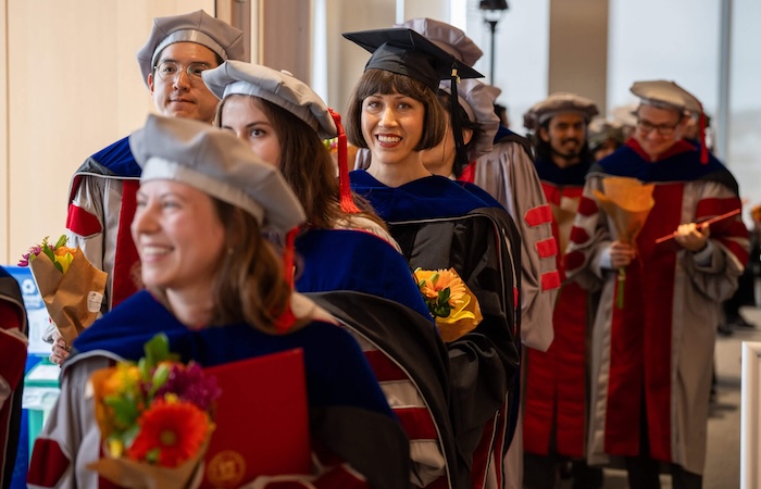 Image of advanced degree graduates processing to their ceremony. They are wearing full regalia and smiling.