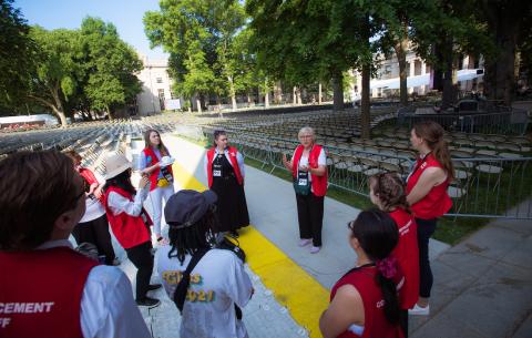 Image of a group of volunteers gathered in a circle receiving instructions. The volunteers are all wearing a red vest that says MIT Commencement on the back
