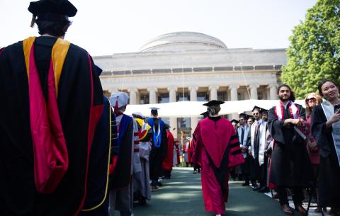 Image of the backs of faculty members in the academic procession. The Killian Court stage and Building 10 are seen in the distance.